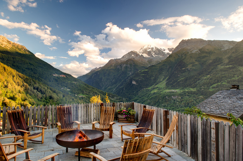 Exterior porch view overlooking the Alps on a bright summer day, with a fire and deck chairs in the